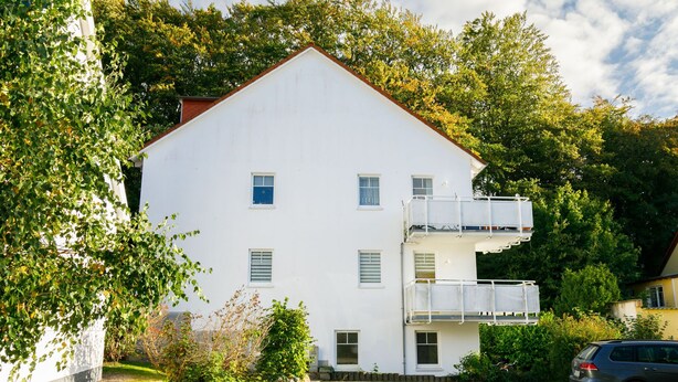 Ein weißes, zweistöckiges Gebäude mit flachem Dach, einem kleinen Balkon im zweiten Stock und einer gepflasterten Auffahrt.