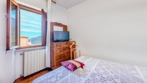 Un dormitorio con un aparador de madera, un televisor encima, una ventana con cortinas y una cama con colcha estampada.