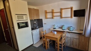 Eine kompakte Küche mit Mikrowelle, Herd, Spüle und kleinem Kühlschrank. Ein Holztisch mit Stühlen steht bereit, und an der Wand ist ein Fernseher angebracht.