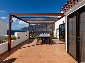 Eine überdachte Terrasse mit Tisch und Stühlen, Blick auf das Meer und ein Berg im Hintergrund.