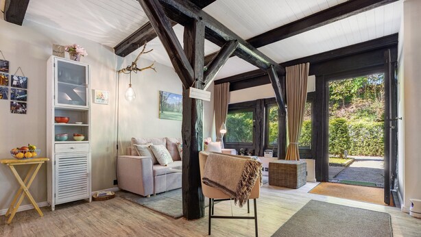 A room with exposed wooden beams, a sofa, a chair with a blanket, a cabinet with dishes, and a view of greenery outside. - Apartment 'Wohnen Im Heidehaus' with Private Terrace and Wi-Fi (St. Dionys)