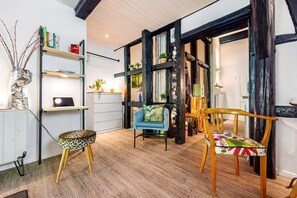 A room with exposed wooden beams, a blue armchair, a wooden chair, a leopard print stool, and a shelf with books and decorative items.