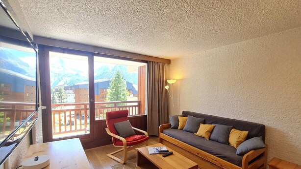 Ein gemütliches Wohnzimmer mit einer Couch, einem roten Sessel, einem Holztisch und einem Balkon mit Bergblick.
