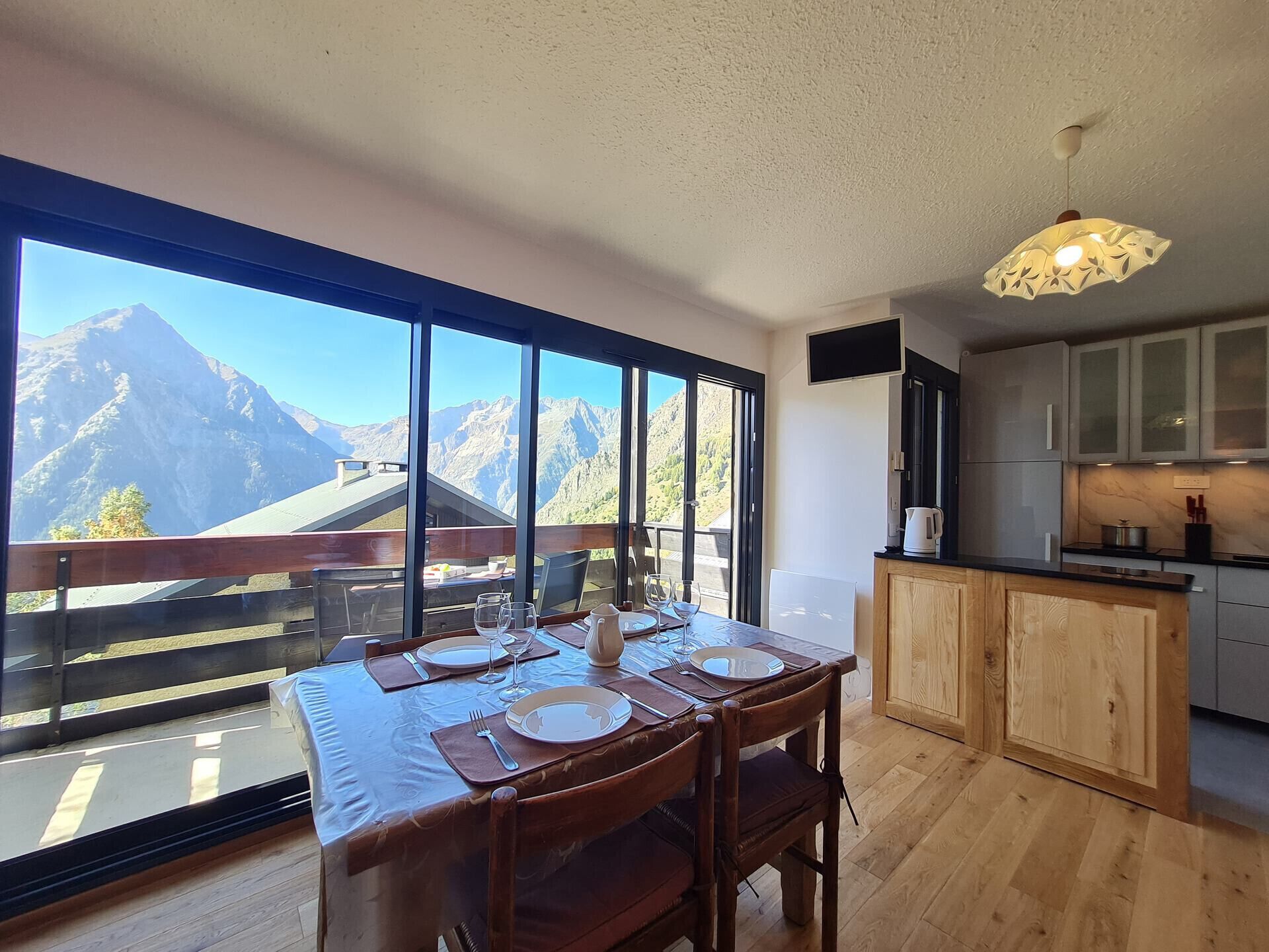 A dining area with a table set for four, large windows offering a mountain view, a kitchen with wooden cabinets, and a hanging light fixture.