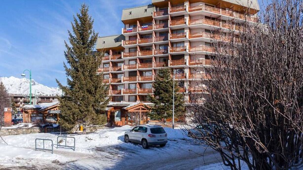 Ein mehrgeschossiges Hotel mit Balkonen, im Hintergrund ein Skigebiet und verschneite Flächen.