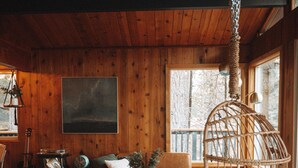 A cozy cabin interior with wooden walls, a hanging chair, a sofa, and a dining area. - Lightfoot Cabin (Running Springs)