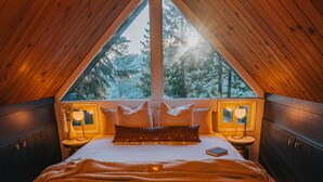 A cozy, triangular-roofed bedroom with a large bed, bedside lamps, and a view of trees through a large window. - Lightfoot Cabin (Running Springs)