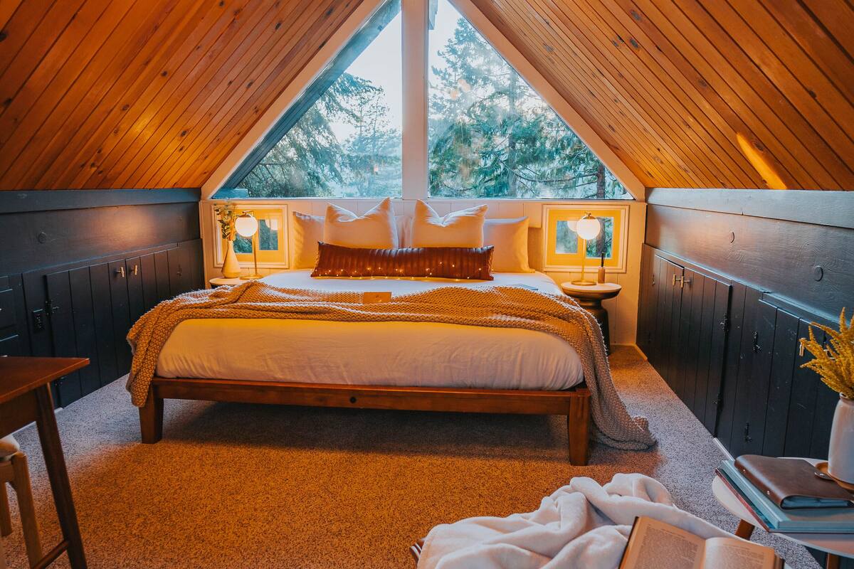 A cozy attic bedroom with a wooden ceiling, a large bed with white bedding, a reading area with a chair and a table, and a view of trees through a window.