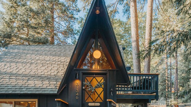 A cabin with a steeply pitched roof, a wooden deck, and a staircase leading to a front door with a wreath. - Lightfoot Cabin (Running Springs)