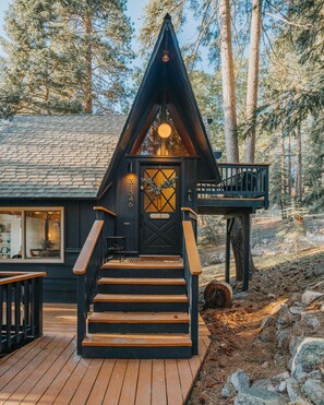 A cabin with a steeply pitched roof, a wooden deck, and a staircase leading to a front door with a wreath. - Lightfoot Cabin (Running Springs)