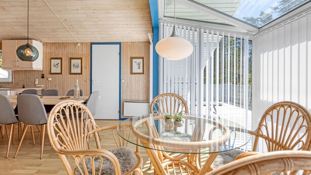 A modern interior with a dining area featuring wicker chairs and a glass table, wooden walls, and a large window providing natural light.