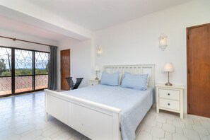 Une chambre à coucher avec un lit, des tables de chevet, une vue à travers une grande fenêtre et un balcon.