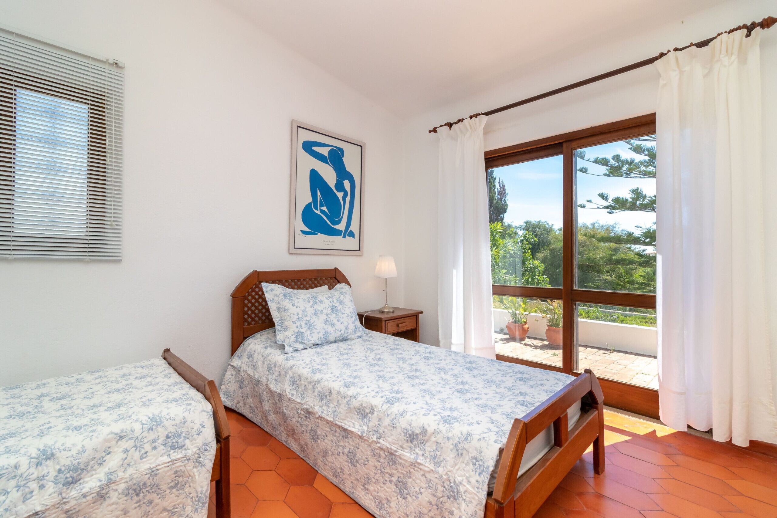 A bedroom with two beds, a window with curtains, a painting on the wall, and a balcony with potted plants.