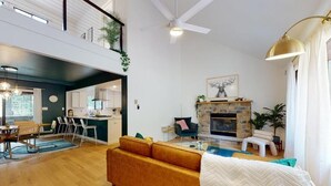 A spacious living area with a high ceiling, a fireplace, and a dining area with a kitchen in the background.