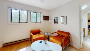 Una habitación con dos sillones de cuero naranja, una mesa de mármol blanco y una mesita auxiliar. Hay tres ventanas con persianas, un ventilador de techo y un cuadro en la pared.