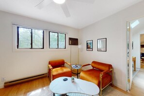Een kamer met twee oranje leren fauteuils, een witte marmeren tafel en een klein bijzettafeltje. Drie ramen met jaloezieën, een plafondventilator en een lijst met een afbeelding aan de muur.