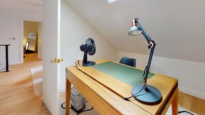 Una habitación con un escritorio de madera, una lámpara de escritorio, un ventilador y vistas a otra habitación con un espejo y una puerta.