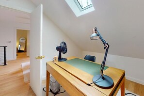 Een kamer met een houten bureau, een bureaulamp, een ventilator en uitzicht op een andere kamer met een spiegel en een deur.