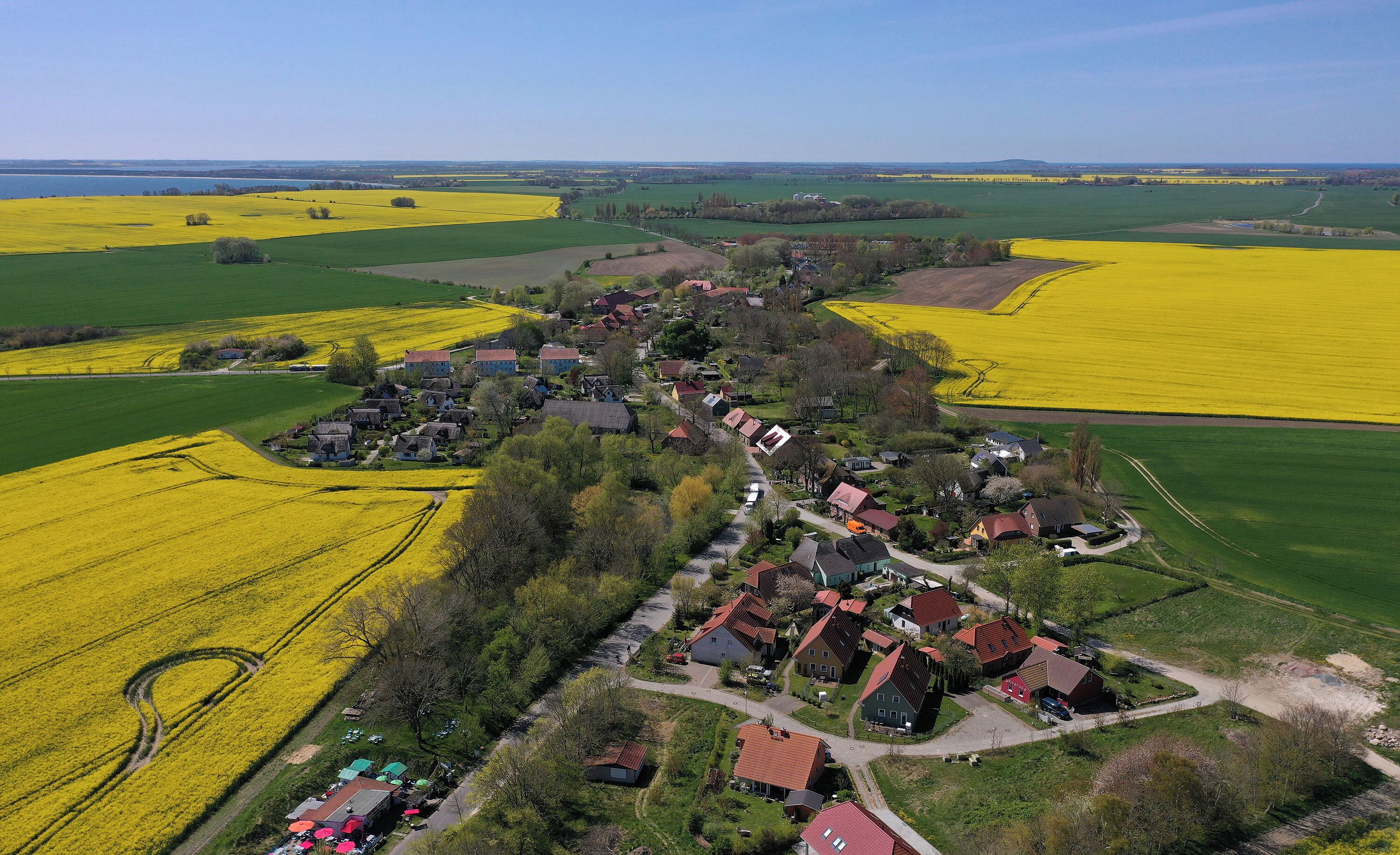 Een vogelvluchtperspectief van een landelijk dorp, met huizen gegroepeerd rond een centrale weg, omringd door velden met gele gewassen en groene velden.