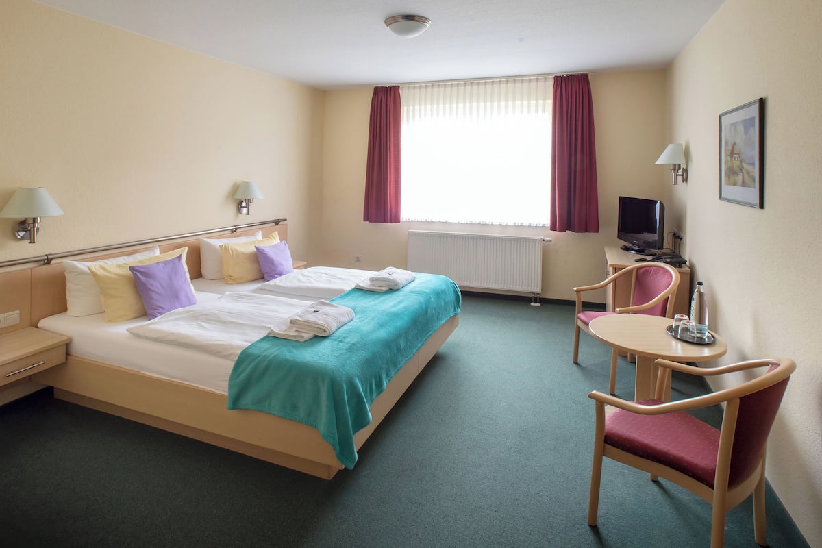 A hotel room with a bed, a desk with a computer, a chair, and a window with curtains.