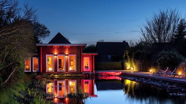 Een huis met een vijver ervoor, een terras met stoelen en een goed verlichte ingang.