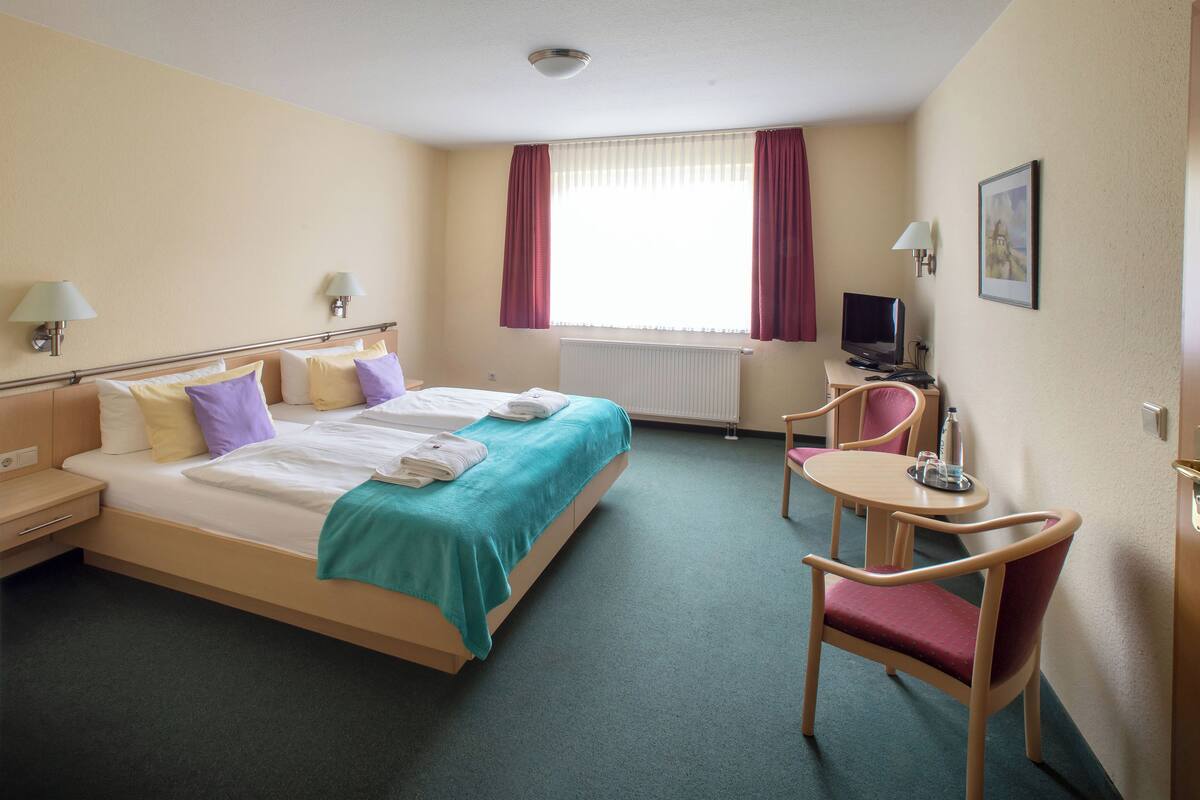 A hotel room with a bed, a desk with a chair, a television, and a window with curtains.