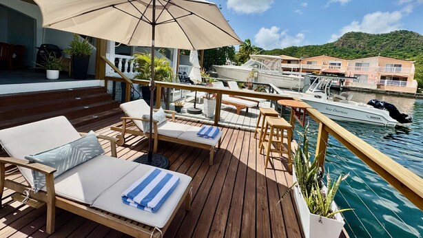 A wooden deck with lounge chairs and a large umbrella, overlooking a marina with boats and a building in the background. - Exquisite Waterfront Villa (Jolly Harbour)