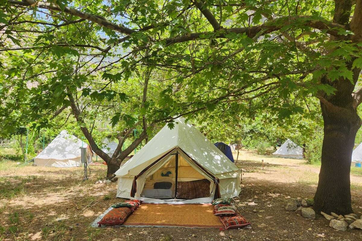 Una tienda de estilo casa árbol con techo inclinado, rodeada de árboles, sobre una zona cubierta de césped con un tapete tejido y cojines en el suelo.