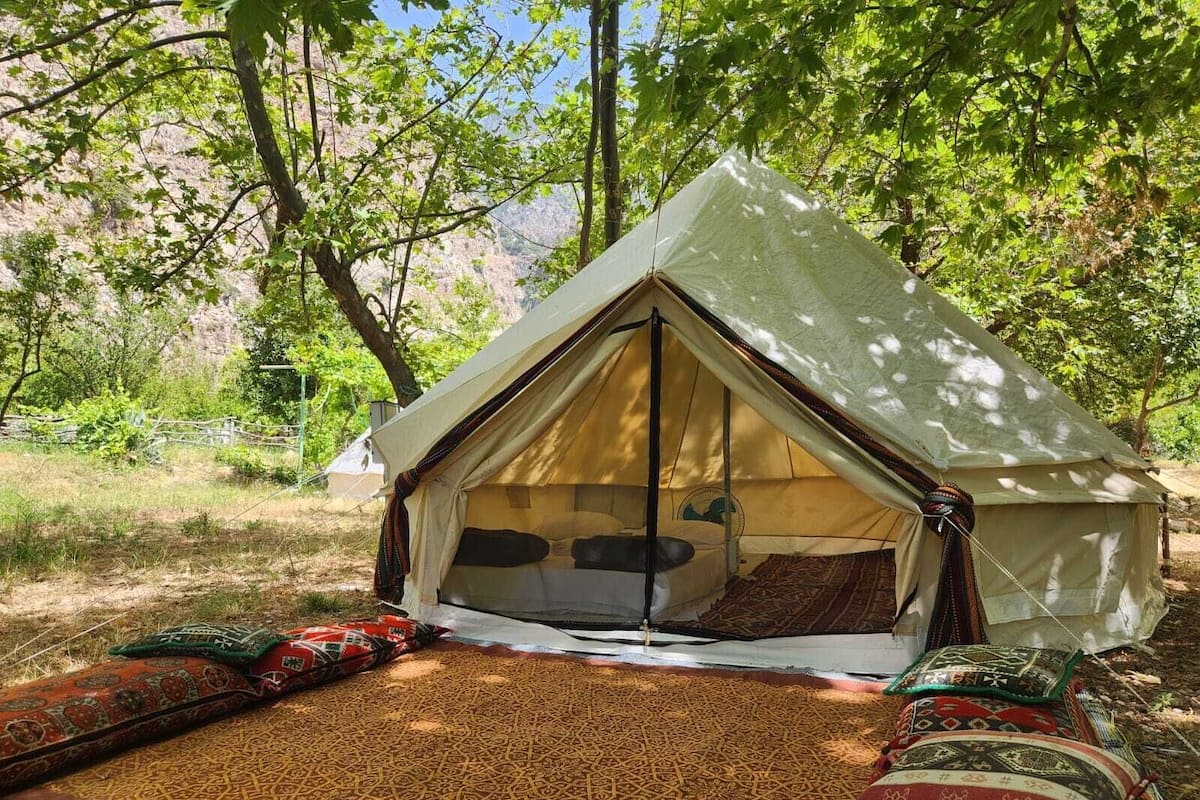 Tienda de campaña montada en un bosque, con una cama en su interior y una manta en el exterior.