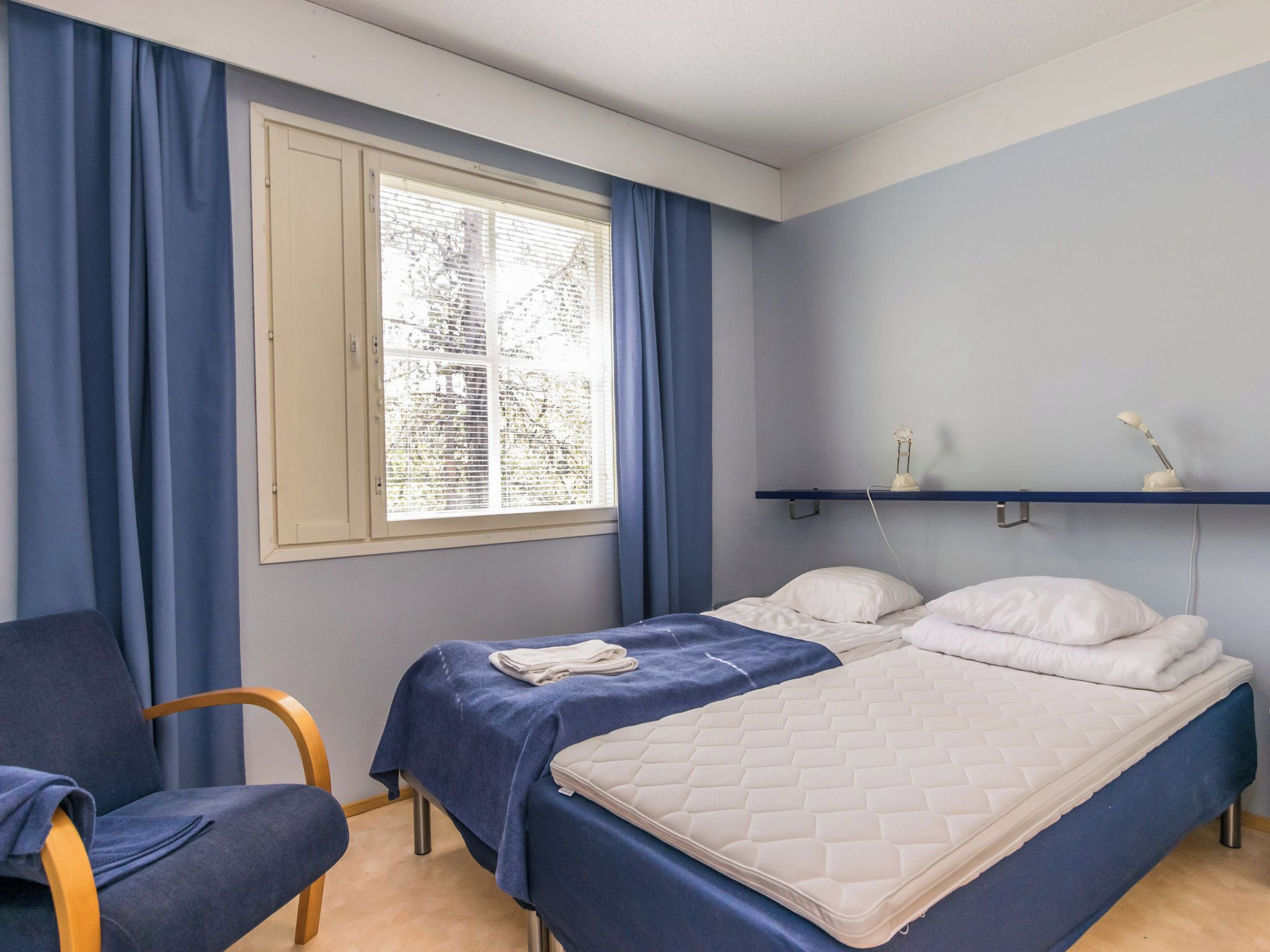 A room with two beds, a chair, a window with curtains, and a bedside table with lamps.