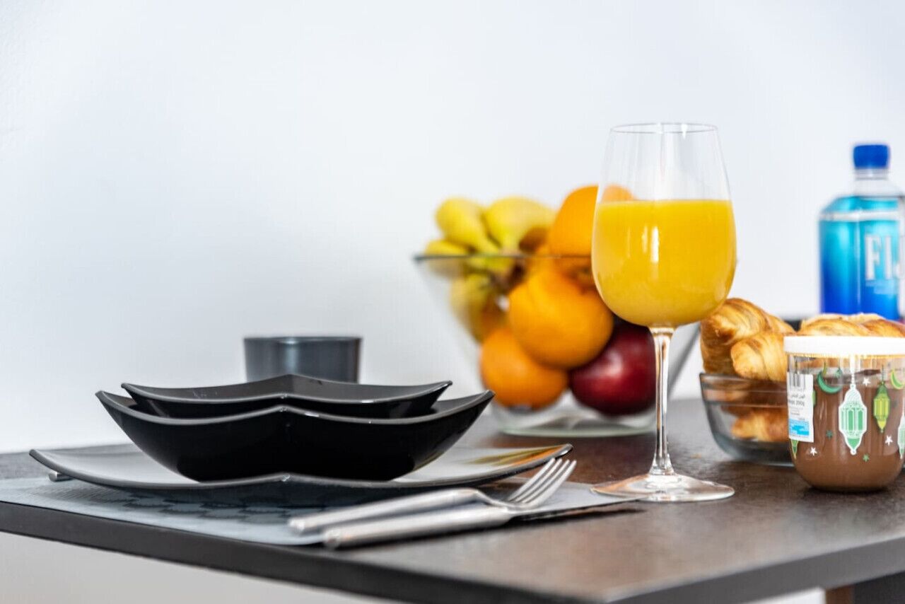 Uma mesa posta para o café da manhã, com um copo de suco de laranja, uma tigela de frutas, croissants e um pote de geleia.