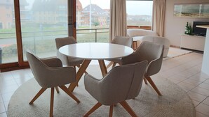 Un comedor moderno con una mesa redonda y seis sillas, ventanas amplias con cortinas y vistas a edificios residenciales.