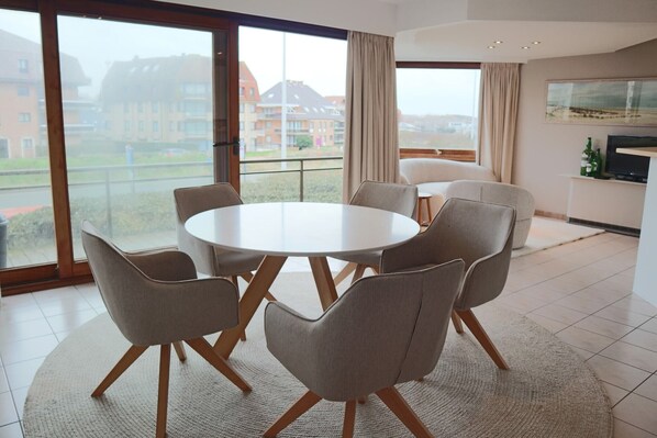 A modern dining area with a round table and six chairs, large windows with curtains, and a view of residential buildings.