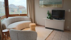 Una sala moderna con un sofá curvo, un taburete de madera, un televisor sobre un soporte blanco y una ventana con vistas a edificios.