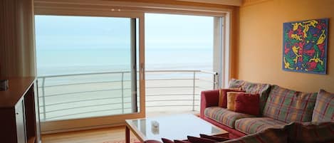 A living room with a sofa, a coffee table, and a view of the sea through a sliding glass door.