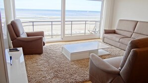 Un salón con un gran ventanal con vistas a una playa, sofás color beige y una mesa de centro blanca.