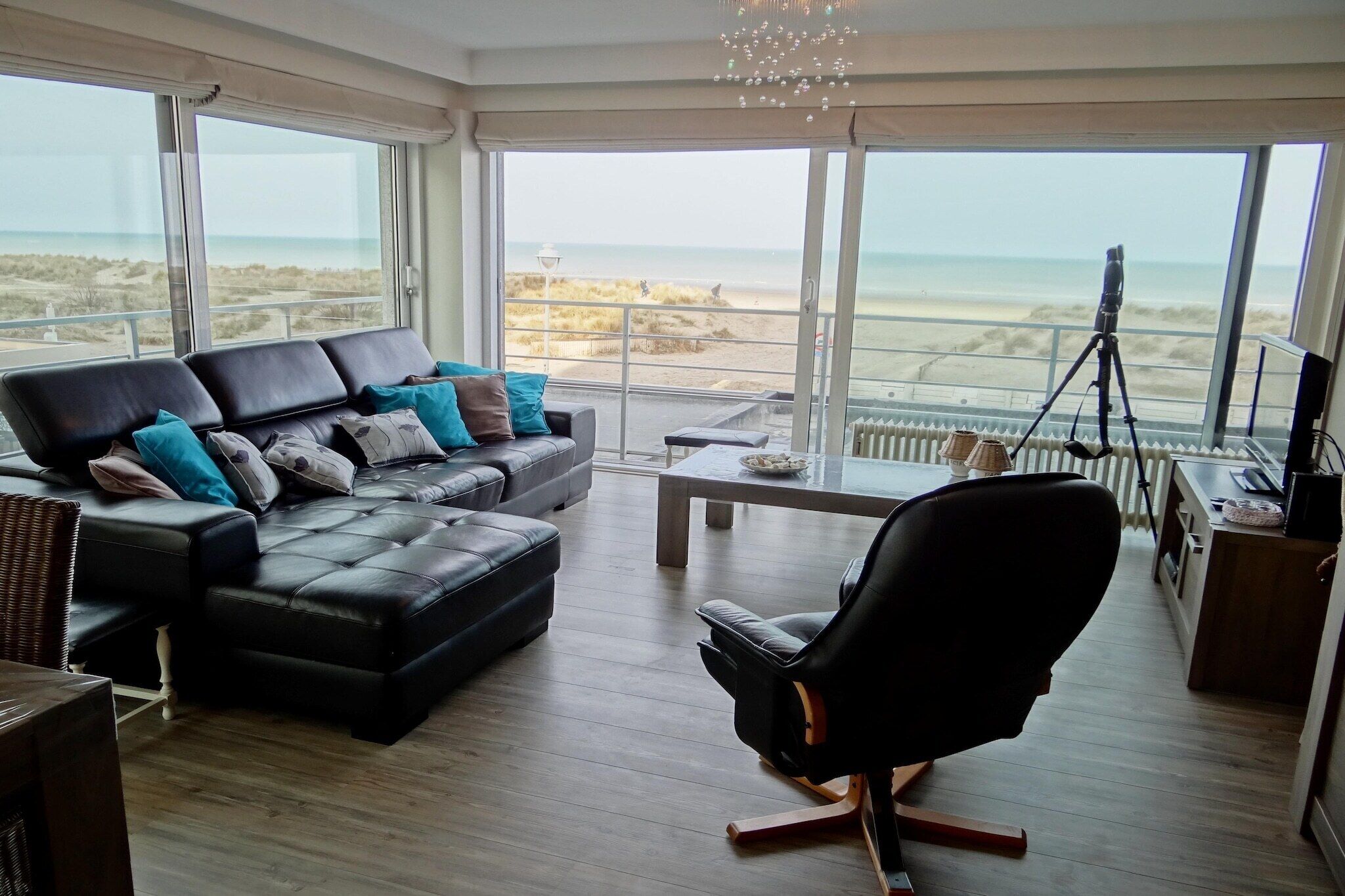A modern living room with a black leather sofa, a black office chair, and a dining table. Large windows offer a view of the beach and ocean.