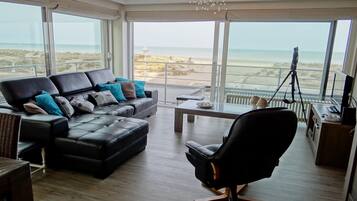 A modern living room with a black leather sofa, a black office chair, and a dining table. Large windows offer a view of the beach and ocean.