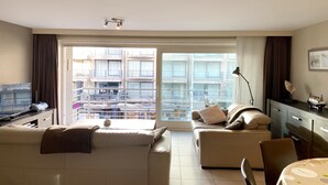 A modern living room with a sofa, a small table, and a TV. There is a large window with a view of buildings and a balcony. - Apartment in Nieuwpoort near the Beach (Nieuwpoort)