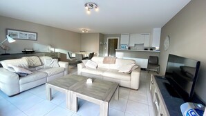 A modern living room with two white sofas, a coffee table, a TV, and a kitchen area in the background. - Apartment in Nieuwpoort near the Beach (Nieuwpoort)