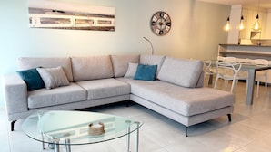 A modern living room with a sectional sofa, a glass coffee table, a wall clock, and a dining area with chairs and a kitchen in the background. - Apartment in Nieuwpoort with Sea Views (Nieuwpoort)