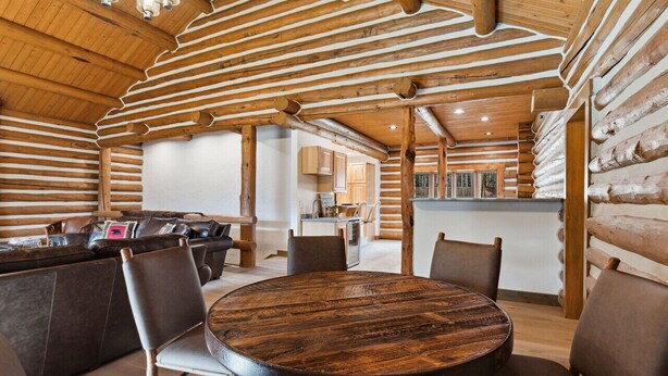 L’intérieur d’une cabane en rondins, avec une table ronde en bois, des sièges en cuir et un coin cuisine visible à travers une porte ouverte.