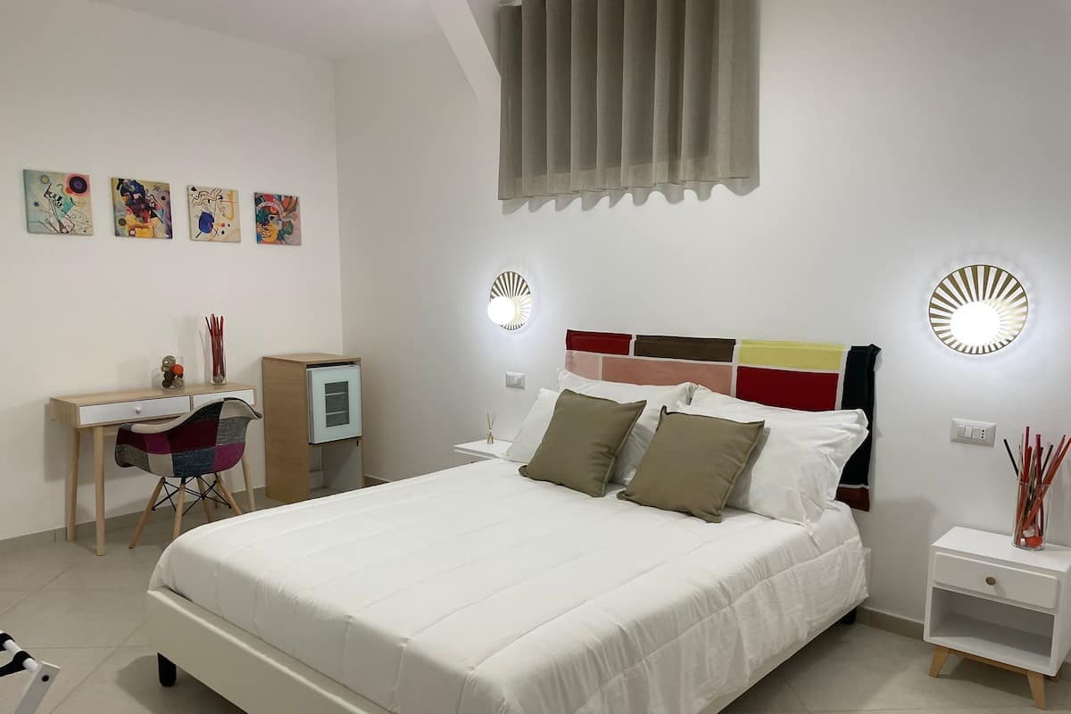 Ein modernes Schlafzimmer mit einem großen Bett, einem Schreibtisch und einem Sessel. An den Wänden sind Wandleuchten und Kunstwerke angebracht.