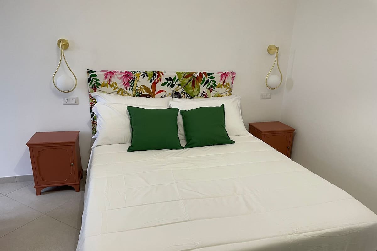 Ein Bett mit weißer Bettwäsche, zwei grünen Kissen und einem Kopfstück mit Blumenmuster. Auf jeder Seite des Bettes befinden sich zwei Nachttische mit Lampen.