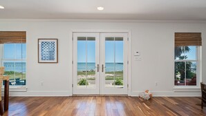Een kamer met houten vloer, een grote glazen deur met uitzicht op het strand, en een ingelijste kunstwerk aan de muur.