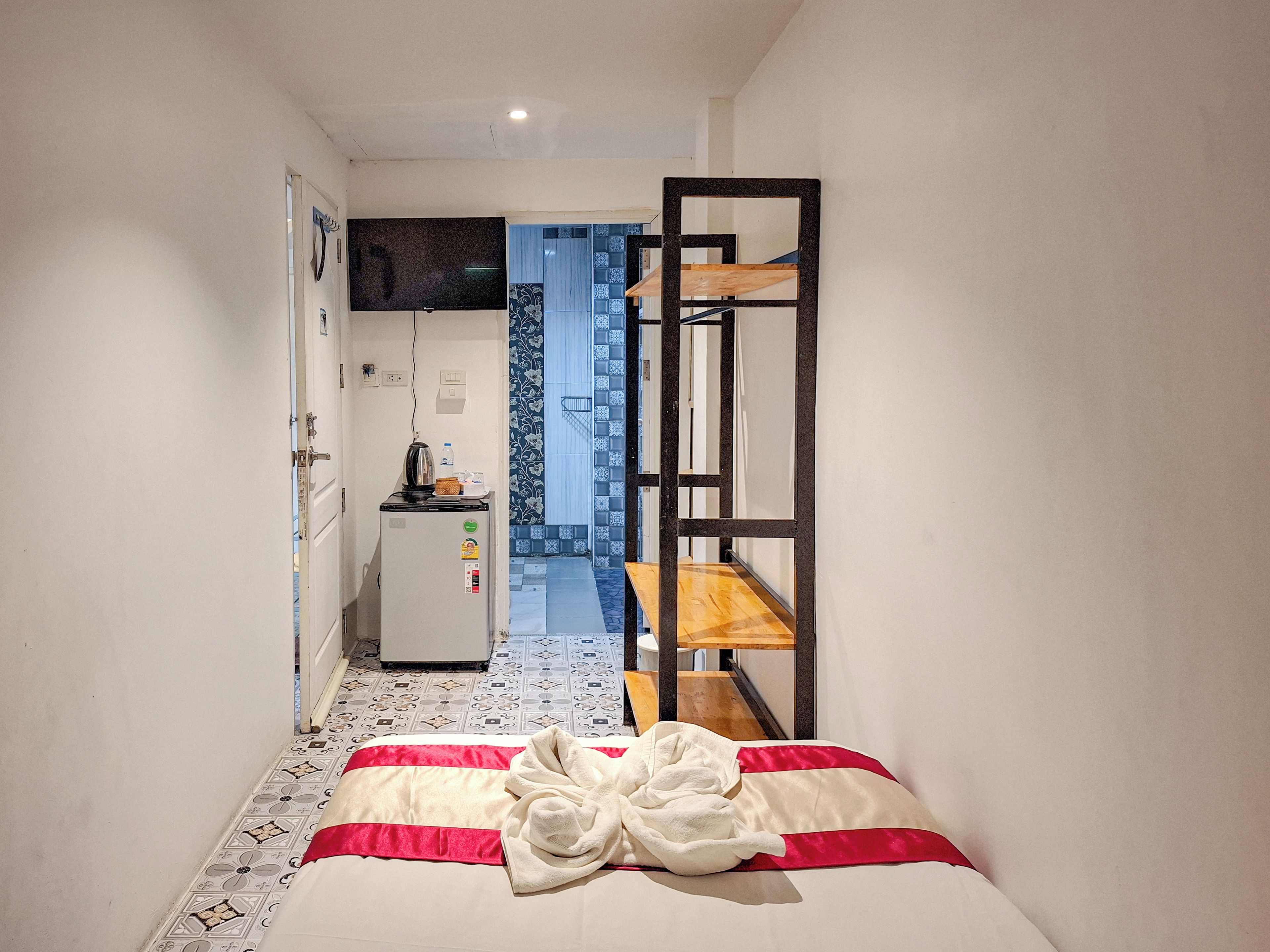 A narrow hotel room with a bed, a small kitchenette, a TV mounted on the wall, and a wooden bunk bed structure.