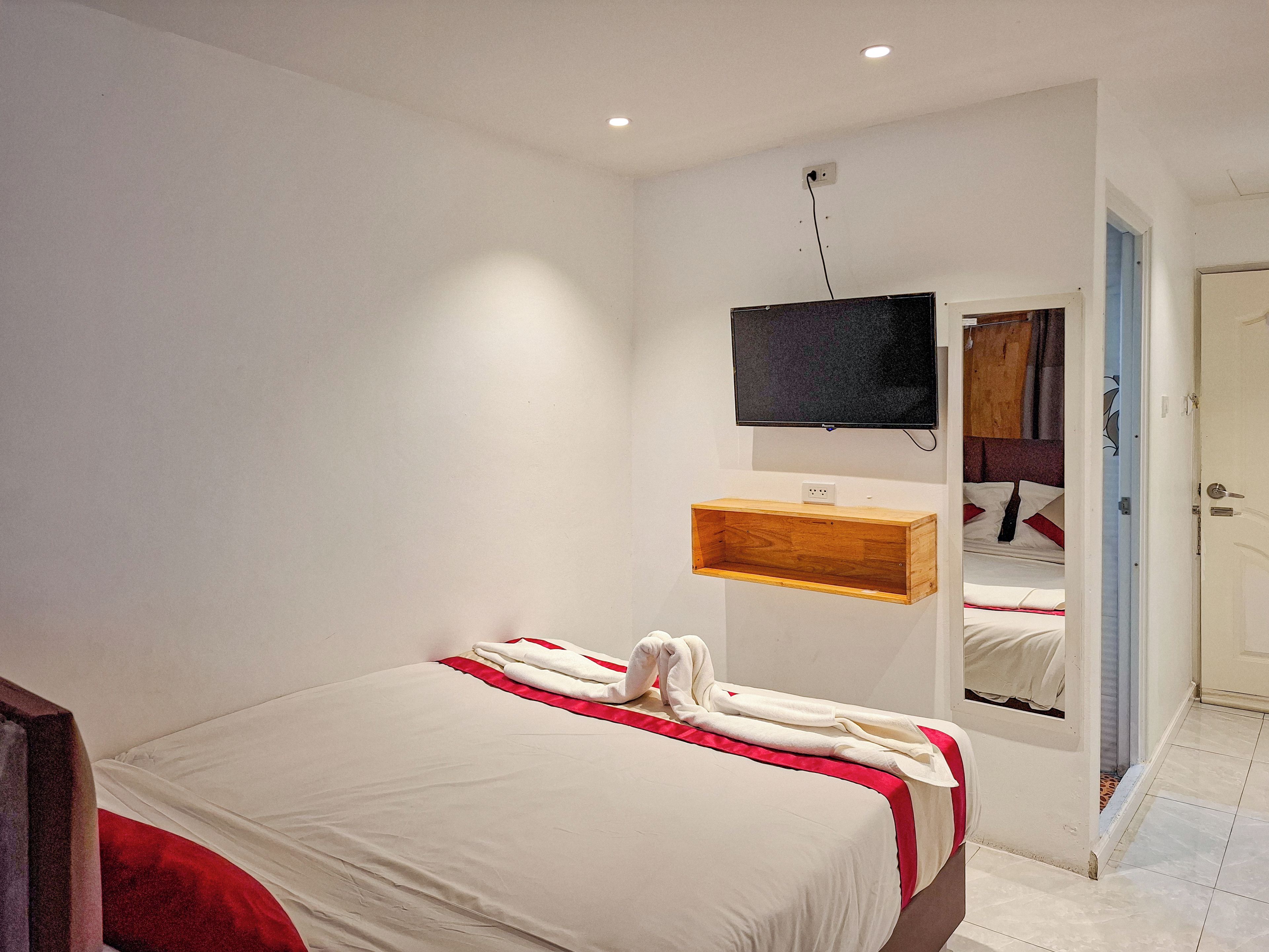Una habitación de hotel con una cama, un televisor montado, un estante de madera y un espejo.
