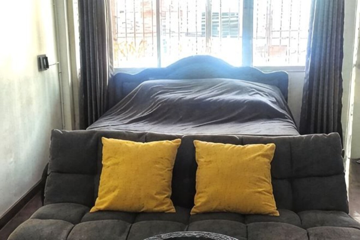 Un lit avec une tête de lit sombre, un canapé gris avec deux coussins jaunes et une fenêtre avec des rideaux.