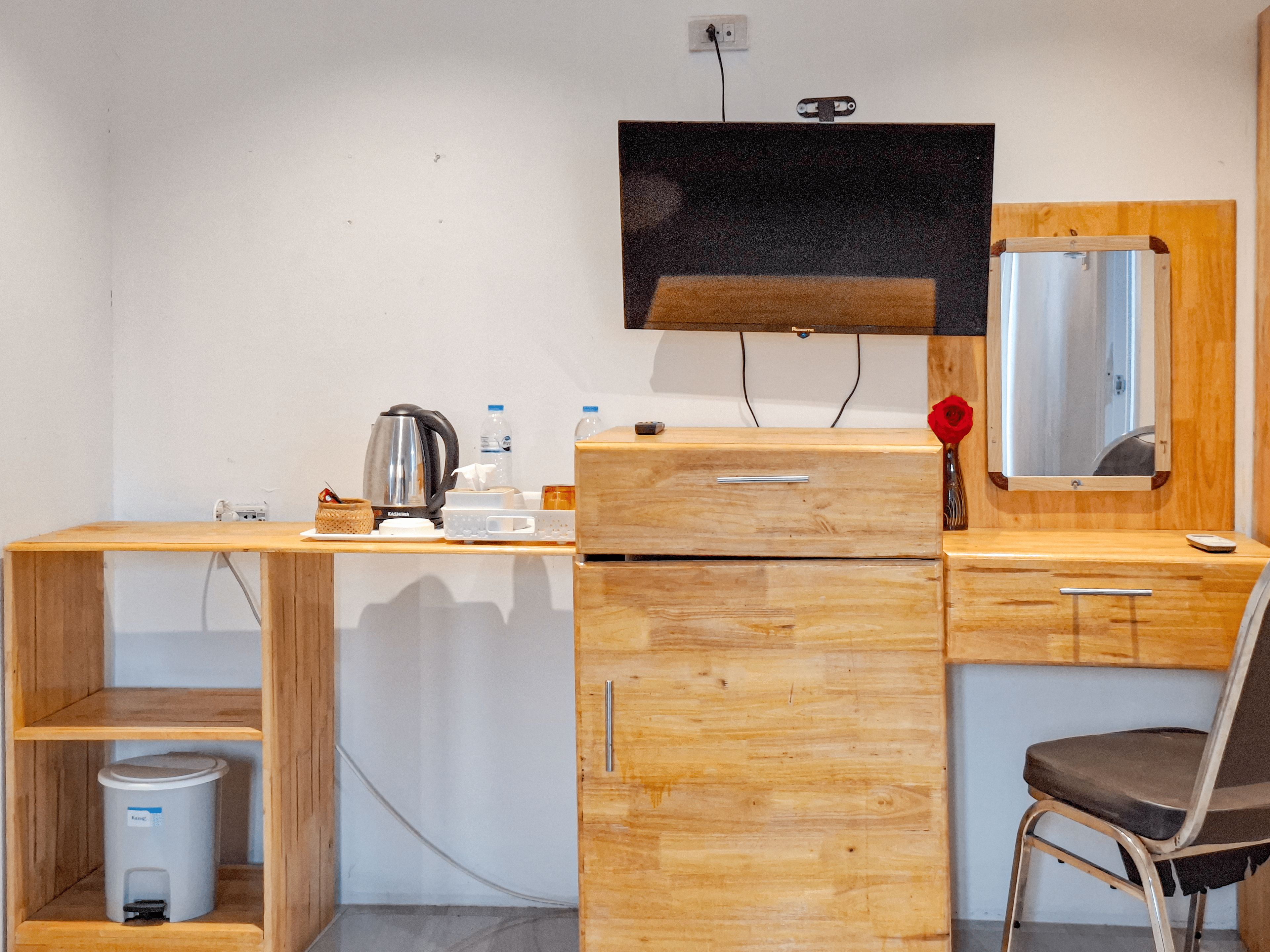 A compact room with a wooden desk, a chair, a television mounted on the wall, a small trash bin, and a mirror.