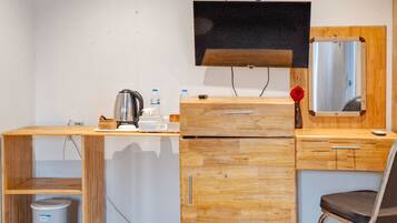 A compact room with a wooden desk, a chair, a television mounted on the wall, a small trash bin, and a mirror.
